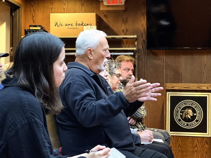 An image of man talking with people beside him on a bench at the Seal of the City of Oneonta New York.